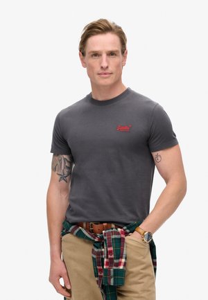 Grijze T-shirt met korte mouwen en een rood logo op de borst, gecombineerd met beige broek en een groen geruit overhemd dat om de taille is gebonden.