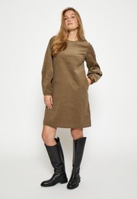 Braunes Cordkleid mit geradem Schnitt, langen Ärmeln und seitlichen Taschen, kombiniert mit schwarzen kniehohen Stiefeln.