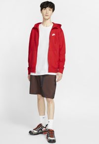 Rode zip-up hoodie met voortassen over een wit T-shirt. Zwart geruite shorts en zwarte sneakers met rode accenten, gecombineerd met witte sokken.
