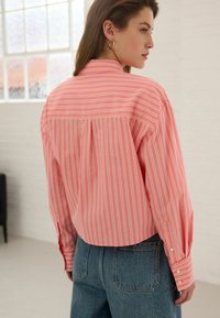 Lichtroze langemouwenshirt met witte en gele verticale strepen, voorzien van een klassieke kraag en opgerolde mouwen. Spijkerbroek zichtbaar eronder.