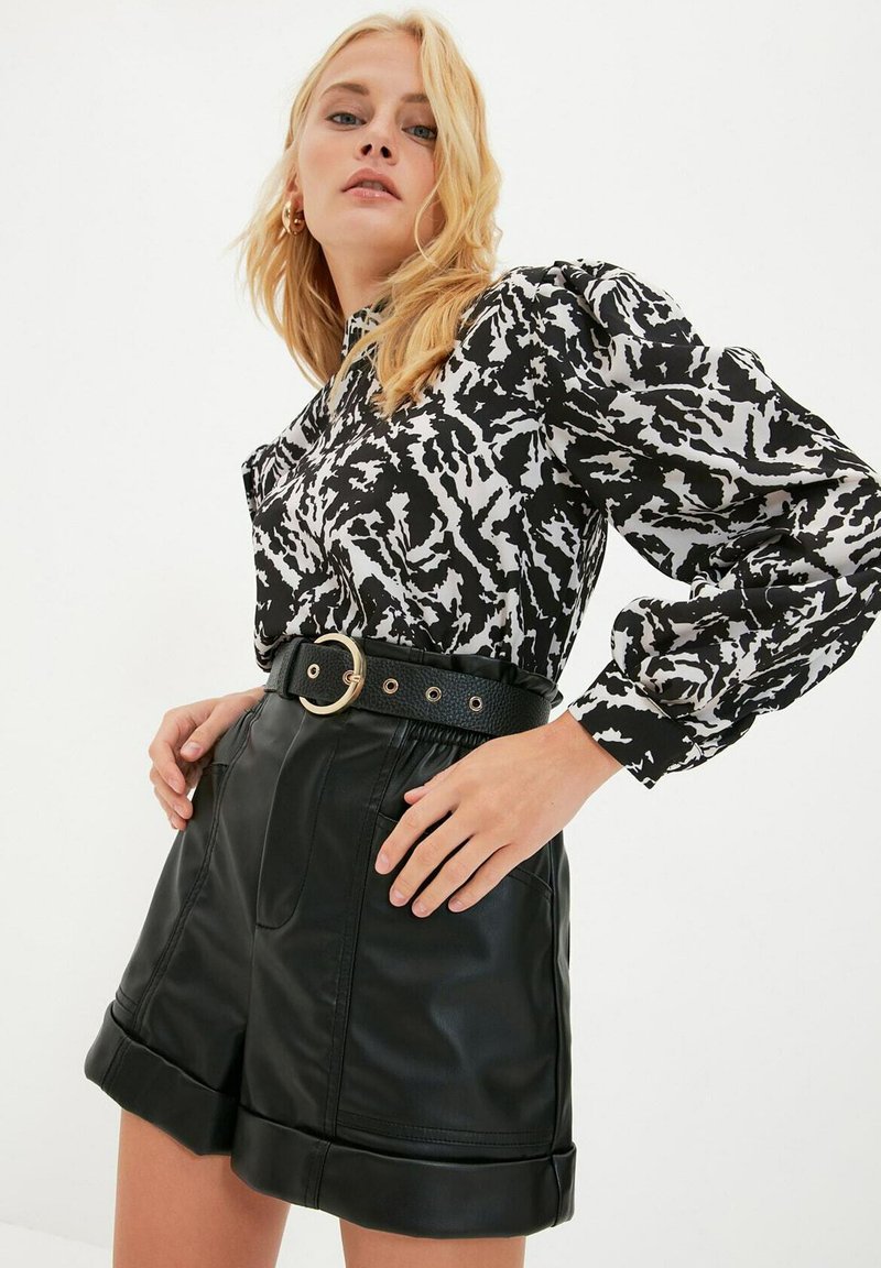 Czarna i biała bluzka w wzory z bufiastymi rękawami, zestawiona z czarnymi szortami z syntetycznej skóry o wysokim stanie oraz szerokim czarnym paskiem z złotą klamrą.