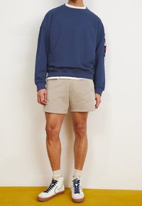 Felpa blu a collo rotondo, pantaloni corti beige, sneakers alte bianche. La felpa presenta una toppa sul petto e le maniche sono leggermente oversize.
