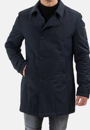 Homme portant un manteau bleu marine foncé à simple boutonnage avec trois boutons, poches latérales et un écusson noir sur la manche gauche, associé à un jean gris.