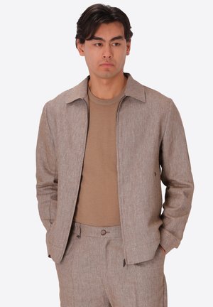 Giovane uomo con capelli scuri in piedi con giacca beige, pantaloni coordinati e camicia marrone chiaro, mani nelle tasche, guarda a destra.