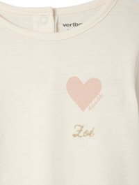 Weißes Baumwollhemd mit Rundhalsausschnitt, das ein hellrosa Herz-Graphic und goldgestickten Text "Zol" darunter zeigt. Mit einem Knopf im Rücken befestigt.