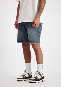 Een persoon in een wit t-shirt en denim shorts staat, met opvallende sneakers en witte sokken, op een neutrale achtergrond.