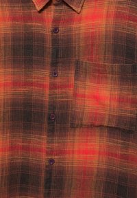 Chemise à carreaux marron, rouge et jaune boutonnée, avec un col et une poche poitrine côté droit.
