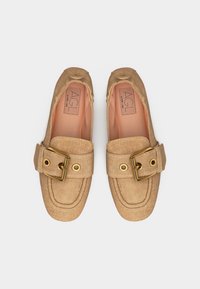 Paire de mocassins en daim beige avec de grandes boucles dorées et des détails cousus, présentée côte à côte sur un fond blanc.