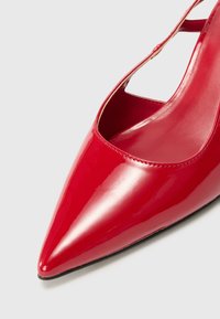 Red patent leather pointed-toe pump with a cut-out side design and a glossy finish. Features a thin ankle strap and a sleek silhouette.