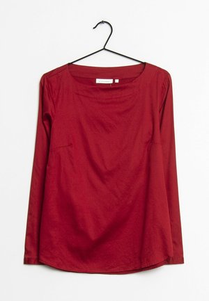 Blouse à manches longues rouge foncé avec col rond, suspendue sur un cintre noir sur fond blanc.