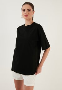 Musta puuvillatee paita, jossa on pyöreä kaula-aukko ja lyhyet hihat, rento istuvuus ja ei näkyviä kuvioita tai yksityiskohtia. Valkoiset shortsit on pidetty.