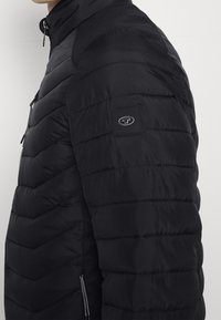 Sort puffer-jakke med lynlås foran, teksturerede vandrette paneler og et logopatch på venstre ærme. Har høj krave og sidelommer.