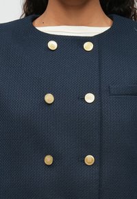 Marinefarget teksturert jakke med dobbeltradet design, som har seks gullknapper og en skjult lomme på brystet.