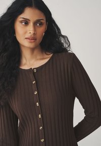 Haut marron à manches longues côtelées avec un col dégagé, présentant une rangée de boutons dorés sur le devant et une silhouette ajustée.