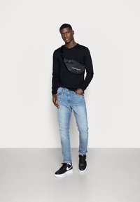 Camisa negra de manga larga, jeans azul claro, zapatillas negras y blancas, y una riñonera negra con un logo, situados sobre un fondo plano.
