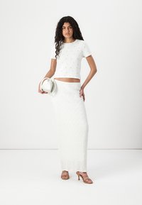 Witte cropped top met kleine gele bloemenprints, gecombineerd met een lange, gestructureerde witte rok; model houdt een kleine witte handtas vast en draagt sleehakken.