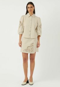 Beige cropped skjortejakke med rund krave og sølvknapper, parret med en matchende nederdel med intrikate broderidetaljer.