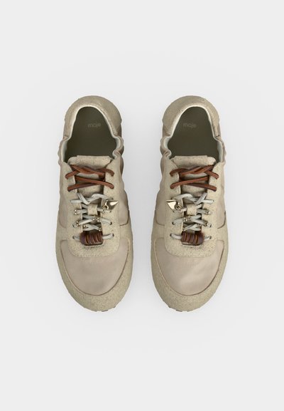 Μπεζ sneakers με υφή, διχρωμικά καφέ και λευκά κορδόνια και μεταλλικά διακοσμητικά κορδονιών, από πάνω σε λευκό φόντο.