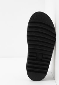 Semelle de chaussure en caoutchouc noir avec des rainures horizontales et des zones texturées au niveau des orteils et du talon, positionnée verticalement sur un fond blanc.