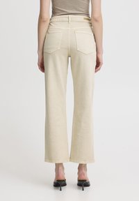 Jeans beige a gamba larga con una texture liscia, due tasche posteriori e una vita piatta. Abbinati a mule con tacco neri.