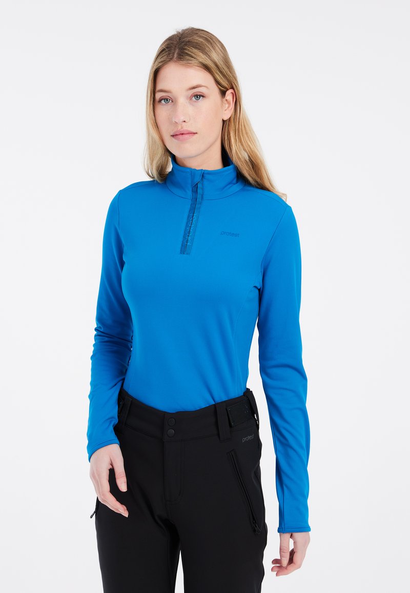 Blå långärmad tröja med hög krage, halvdragkedja, slät yta, och svarta byxor med bälte och fickor med dragkedja.