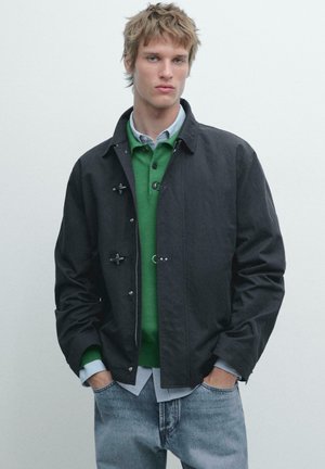 Mladý muž s rozcuchanými blond vlasy, oblečený v černé bundě přes zelený svetr a světle modrou košili, s rukama v kapsách modrých džínů.