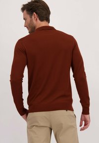 Bruine nauwsluitende trui met lange mouwen en een gladde textuur, geribbelde manchetten en zoom. Model is iets gedraaid om het ontwerp van de achterkant te tonen tegen een neutrale achtergrond.