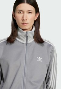 Siva zip-up jakna s visokim ovratnikom, s tri bijele pruge na svakom ramenu i malim bijelim Adidas logotipom na prsima.