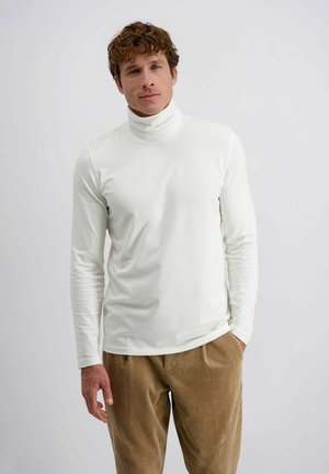 Man in een witte coltrui met lange mouwen en een bruine corduroy broek, staand tegen een effen lichte achtergrond.