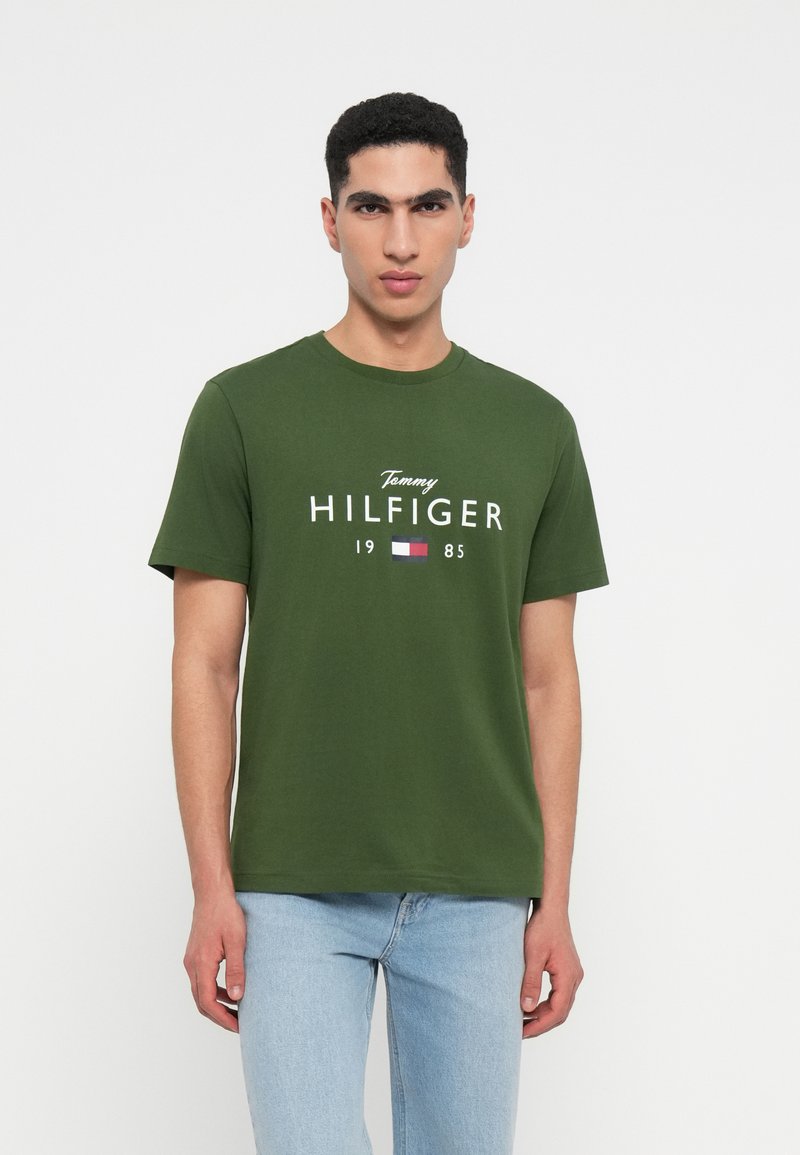 Zelené bavlněné tričko s bílým textem a logem "Tommy HILFIGER" a nápisem "1985" pod ním. Kulatý výstřih a krátké rukávy.