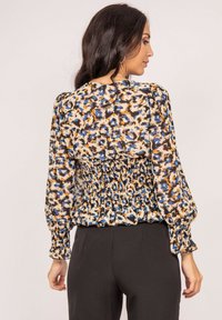 Blouse à dos smocké en tissu beige avec un motif abstrait bleu, orange et noir, présentant de longues manches bouffantes et une taille froncée.