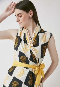 Wrap dress featuring black and white fabric with an abstract floral pattern in yellow. Accented with a yellow belt tied at the waist.