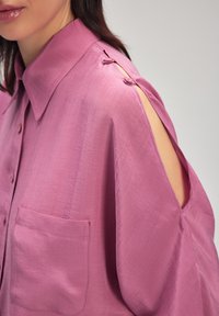 Camisetta rosa, morbida e ampia, con colletto, taglio sulla spalla sinistra, dettaglio con bottoni laterali e taschino frontale. Tessuto liscio e texturizzato.