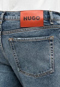 Μπλε τζιν παντελόνι με κόκκινο δερμάτινο ταμπελάκι με ανάγλυφο λογότυπο "HUGO". Περιλαμβάνει τσέπη στην πίσω πλευρά και πορτοκαλί ραφές λεπτομέρειας.