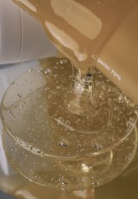 Przezroczysta szklanka wylewająca lepki, jasnobrązowy płyn z bąbelkami, lądujący w lustrzanej szklanej misce, odsłaniający gładkie powierzchnie.