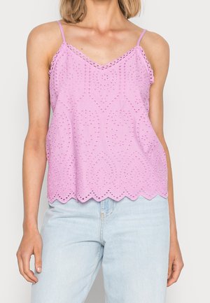Kobieta ubrana w bezrękawnikowy, liliowy top z ażurowej koronki z falowanym dołem oraz jasnoniebieskie, wysokim stanem jeansy na jednolitym tle.