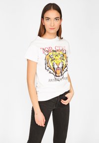 Ung kvinde iført en hvid T-shirt med et brølende tiger grafik og "TOP GUN" tekst, parret med sorte jeans, poserende mod en hvid baggrund.