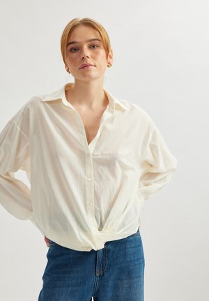 Chemise blanche légèrement ondulée avec une coupe décontractée, manches longues et une poche poitrine, nouée à la taille sur un jean bleu.