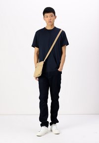 Tricou scurt cu mânecă, de culoare bleumarin, blugi închis și adidași albi. O geantă tip crossbody, de culoare bej, atârnă de umăr, subliniind forma sa rectangulară.