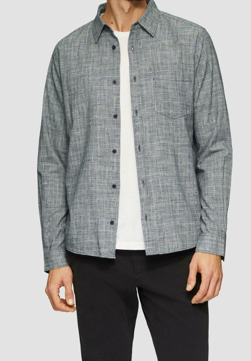 Homme portant une chemise à carreaux grise ouverte sur un t-shirt blanc et un pantalon noir sur un fond uni.