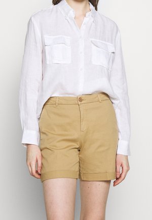 Weißes Leinenhemd mit Knopfleiste und zwei Brusttaschen, kombiniert mit hellbraunen Baumwollshorts. Die Shorts haben einen umgeschlagenen Saum und einen Knopfverschluss.