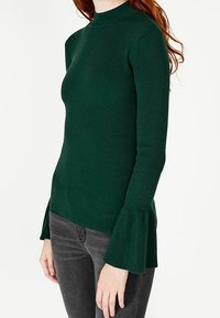 Pull en maille côtelée vert foncé avec un col montant et des manches évasées, présentant une silhouette ajustée et une texture douce.