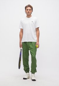 Νεαρός άνδρας που στέκεται κρατώντας ρακέτα του τένις και μπάλα, φοράει λευκό μπλουζάκι, πράσινο παντελόνι και λευκά αθλητικά παπούτσια, μπροστά σε απλό φόντο.