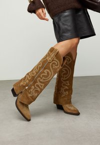 Bottes hautes en daim marron avec une broderie claire complexe, bout pointu et talon bloc. Dotées d'une fermeture éclair latérale pour un enfilage facile.