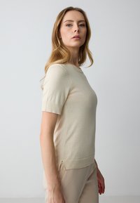 Maglione corto beige a maniche corte con scollo rotondo, polsini e orlo a coste, e una texture liscia. Spacchi laterali vicino all'orlo.
