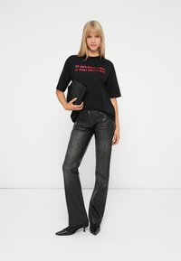 Tricou oversized negru cu text roșu, asortat cu blugi negri mulați și tocuri negre ascuțite. Modelul ține o poșetă neagră.