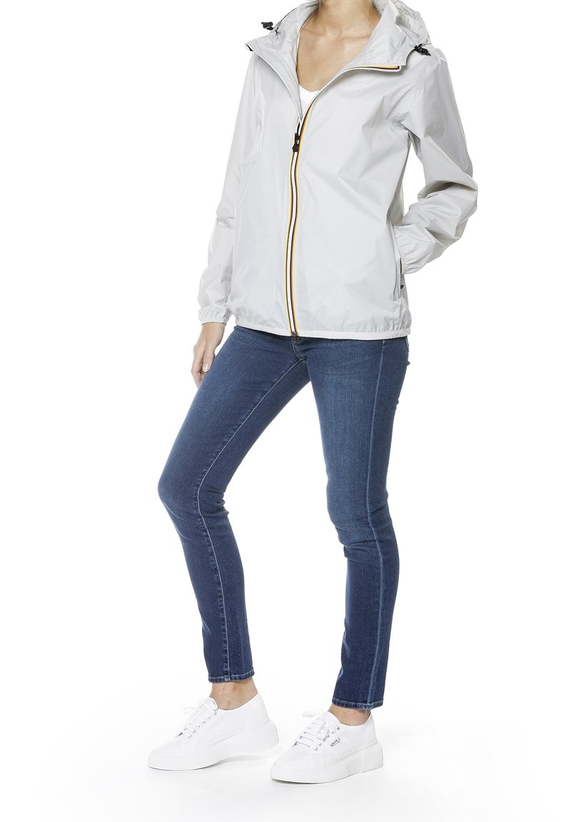 Femme portant une veste à capuche gris clair avec fermeture éclair à l'avant, jean slim bleu et baskets blanches, debout avec une main dans la poche.