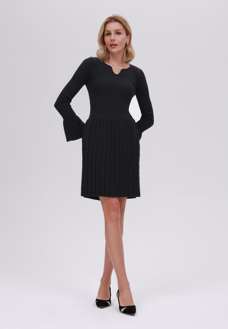 Schwarzes geripptes Kleid mit schwingendem Ärmel, tailliertem Oberteil und plissiertem Rock. Verfügt über einen gewellten Ausschnitt und einen knielangen Schnitt.