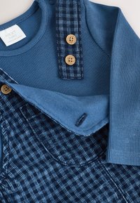 Niebieski prążkowany długi rękaw dla niemowląt z granatowymi, kraciastymi ogrodniczkami zapinanymi na dwa drewniane guziki oznaczone napisem „NEXT”.