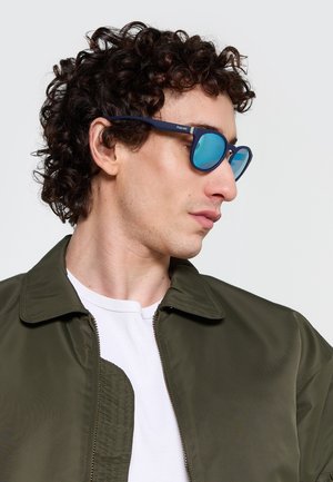 Uomo con capelli ricci e scuri, che indossa occhiali da sole specchiati blu, una camicia bianca e una giacca verde oliva, che guarda di lato.